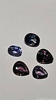 Photo of five natural sapphires in oval and pear cuts showing purple and blue colors with high clarity, laid on a light background.
