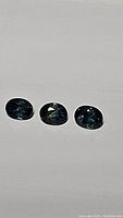 Three oval-shaped natural sapphires laid out on plain surface showing deep blue color with visible internal multicolored reflections.