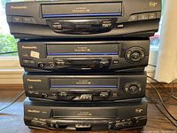 Four black VCR units stacked, three Panasonic and one Quasar model. Panasonic units have 4 Head Division S logo and visible model labels. Quasar unit is black with similar front loading slot.