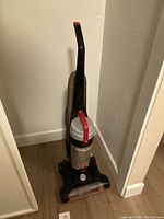 Front view of the black upright vacuum with attached vacuum hose resting against a white wall on wood flooring.