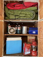 Top wooden shelf holding three sleeping bags: two green and one red rolled up and stacked.