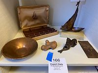 Overview of copper collectibles lot on shelf showing all items described in the lot. Includes catch-all dish, dog sculpture, ship sculpture on alabaster base, copper floral art plate, Hickok men's jewelry box with ship emblem inside lid, and small copper-colored abstract metal piece.
