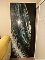 Front view of large painting showing ocean wave on canvas leaning against wall next to furniture.