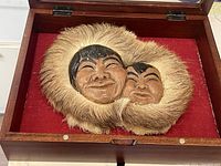 Close-up of the handmade carved and painted Eskimo mother and child faces with fur trim displayed inside a wooden box with red fabric background.