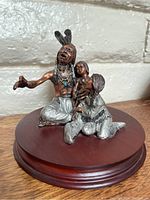 Photo of the full figurine showing three Native American figures seated on a wooden base against a white brick wall backdrop.