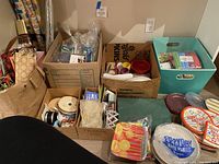 Photo shows multiple boxes and bins filled with various party supplies including paper plates, napkins, ribbons, gift bags, and wrapping paper rolls.