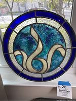 Front view of round stained glass piece with blue border and cream swirl design in center, sitting on a windowsill with sunlight shining through.