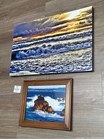 Photo of two ocean scene photographs on floor; one framed print with wooden frame and ocean with rocks, one large mounted print on board with ocean waves and sunset sky