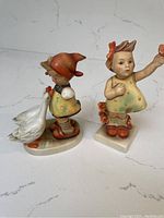 Two vintage Hummel figurines, one child with a goose, the other girl holding hand up, both hand-painted porcelain.