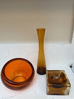 Photo showing three vintage amber glass pieces: a rust-orange bowl, a tall orange-to-dark amber vase, and a square amber ashtray with chips.