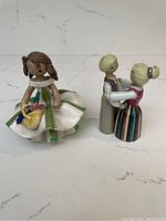 Both vintage ceramic figurines shown together on white background, girl in dress with basket and couple in traditional attire