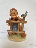 Full front view of the vintage Hummel figurine showing the child peeking over a wooden fence with a blue bird perched on top.
