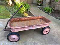Full side view of vintage rectangular red metal wagon with handle, showing rust and weathering from outdoor use.