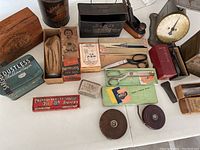 Wide photo showing layout of assorted items on a white surface, various vintage tins, scissors, tape measures, kitchen scale, wooden box, and small tools visible.