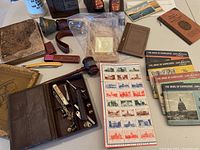 Wide angle of all collectible items arranged on table including antique books, shaving kit, stamps, bronze baby shoes, wooden gavel, stein, postcards and knife