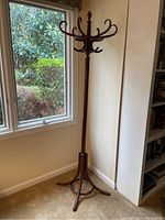 Full view of the wooden coat stand near a window showing the tall height and curved legs of the tripod base.