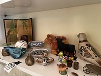 Full view of shelf with assorted collectibles and decor, showing trinket box, pottery dishes, animal figurines including plush camels, black bull, ceramic bird, and others.