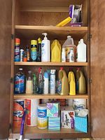 Household cleaning supplies neatly organized inside a wooden cabinet with three shelves containing spray bottles, liquid containers, wipes, tissues, and paper towels.