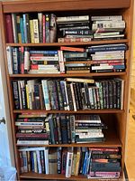 Wooden bookshelves filled with a large assortment of books in various genres and formats. Shows tightly packed books with visible titles from fiction and nonfiction categories.