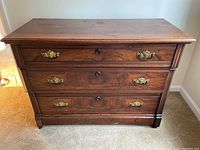 Front view of antique mahogany chest of drawers showing three drawers with ornate brass handles and keyholes, flame mahogany veneer.