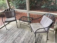 Pair of black metal chairs with grey cushions flanking a small black metal framed glass top table, all placed on a grey area rug inside a screened porch with a view of mulch and bushes outside.
