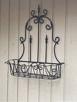 Front view of black metal hanging planter attached to outdoor vertical wood siding showing decorative scrolls and basket base