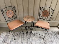 Pair of black metal folding chairs with lattice seats and terra-cotta cushions, flanking small round table. Chairs and table have terra-cotta color decorative tile inserts with leaf patterns.