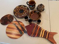 Full view of brown pottery set on table including plate, pitcher, sugar bowl, bowl, and small cups, alongside two wooden trivets shaped round and fish-shaped, and steel carving fork with sharpener.