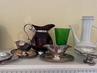 Wide front view of silver plated gravy boats with plates, purple glass pitcher with silver handle, green glass vase, and white large pitcher on mantle.