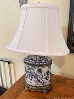 Full view of one blue and white toile table lamp on wood surface with white fabric shade and ornate brass base.