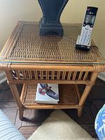 Side view of one wicker side table showing woven wicker top with glass insert, vertical wicker rod detail on legs, and lower wicker shelf with book on it.