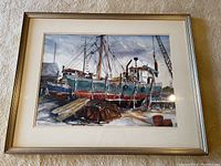 Framed watercolor painting showing fishing boats docked in a harbor with equipment and crane, signed Margaret Laurie.