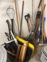 Full view of wheelbarrow with assorted yard tools leaning in corner, showing shovels, rakes, clippers, pruning shears, axe, and wooden yard sticks.