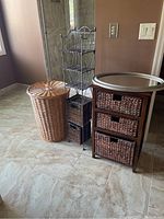 Photo showing a tall beige wicker hamper, narrow black metal storage shelf with three wicker baskets, wooden storage shelf with wicker baskets, and silver metal oval mirror on top in a master bathroom.