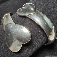 Close-up front view of silver open cuff bracelet with two heart-shaped ends against black background showing polished surface and small scratches.