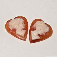 Two heart-shaped cameo pieces each featuring a carved female profile in white on orange background, facing opposite directions.