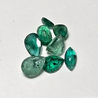 Seven loose emerald gemstones photographed on a white background showing different angles and facets, highlighting the natural inclusions and vibrant green color.
