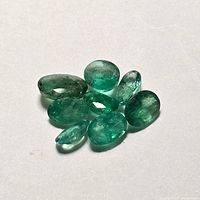 Eight loose emerald gemstones assorted on a white surface, showing varying oval and rounded shapes with translucent green color and inclusions.