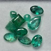 Top view of ten loose emerald gemstones showing varied shapes and green hues on a white felt surface
