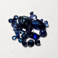 Loose sapphire gemstones arranged on a white surface showing various cuts including round, oval, marquise, pear, and emerald shapes with deep blue color.