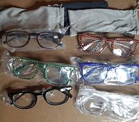 Six pairs of reading glasses in plastic wrap with three glasses bags on a surface. Variety of frame colors and patterns visible.