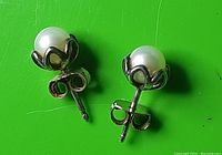Clear close-up photo showing the pair of small stud earrings with pearl beads in a silver floral setting resting on a green background.