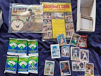 Photo of Baseball Card Collecting Kit booklet, Lumpy baseball book, five individual sealed packs of 1990 Pre-Rookie Baseball Cards, multiple loose baseball cards featuring various players including Astros, and a box with numerous additional cards.