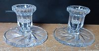 Two clear crystal candle holders with round bases and textured candle sockets, placed on a wooden surface against a dark background.