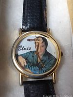 Close-up front view of the Elvis Presley wristwatch showing the gold-tone case, clear face with Elvis image and guitar second hand, and worn black leather strap.