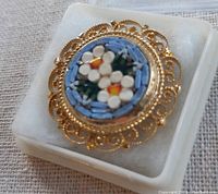 Front close-up view of the Italian mosaic brooch showing the blue, white, green, and orange stone floral pattern surrounded by gold-tone filigree setting, displayed on a white cushion in a box.