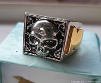 Front view showing the square face with raised silver skull on black background and corner scroll detailing.