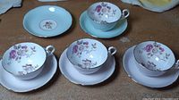 Four teacup and saucer sets arranged with visible floral patterns inside cups, and pink-tinted and blue-tinted saucers with gold edge trim.