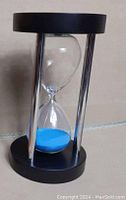 Hourglass with blue sand held by three chrome pillars between black top and base, shown as a demo unit to represent item for sale new in box.