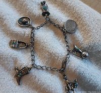 Silver-toned charm bracelet laid flat on light fabric background. Seven distinct western-style charms with turquoise stones visible.